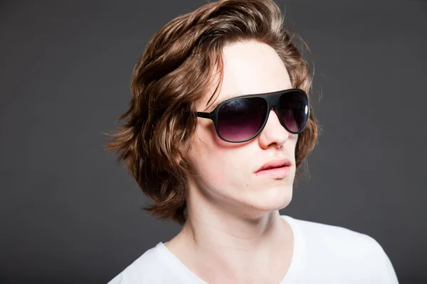 Schöner junger Mann mit braunen langen Haaren und Sonnenbrille auf grauem Hintergrund. Modestudio erschossen. — Stockfoto