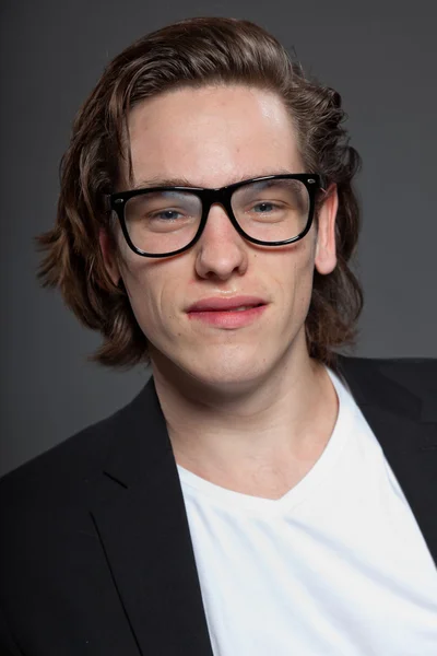 Schöner junger Mann mit braunen langen Haaren und weißem Hemd, blauer Jacke und Retro-Brille auf grauem Hintergrund. Modestudio gedreht. Gesichtsausdruck. — Stockfoto