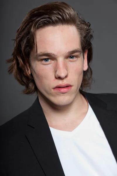 Handsome young man with brown long hair and wearing white shirt and blue jacket isolated on grey background. Fashion studio shot. Expressive face. — Stock Photo, Image