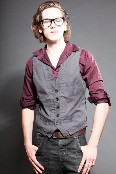 Schöner junger Mann mit braunen langen Haaren auf grauem Hintergrund. trägt eine schwarze Retro-Brille. Modestudio gedreht. Gesichtsausdruck. — Stockfoto