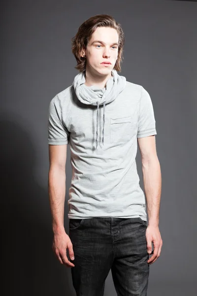 Bello giovanotto dai capelli castani lunghi che indossa una camicia grigia isolata su sfondo grigio. Scatto dello studio di moda. Viso espressivo . — Foto Stock