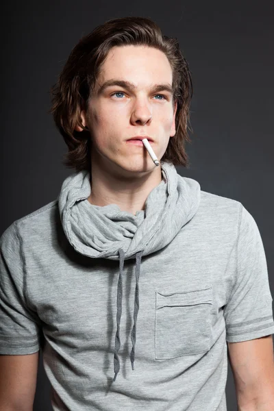 Schöner junger Mann mit braunen langen Haaren, graues Hemd auf grauem Hintergrund. Zigarette rauchend. Modestudio gedreht. Gesichtsausdruck. — Stockfoto