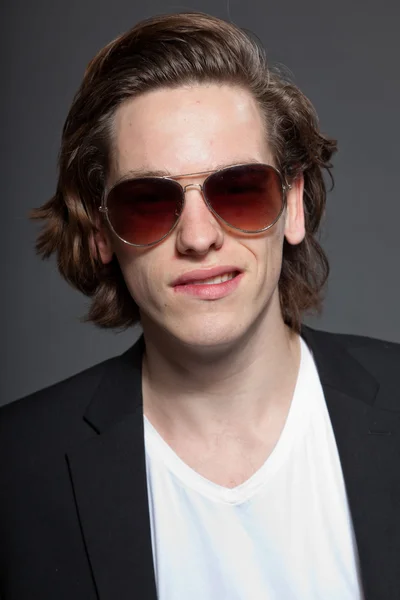 Handsome young man with brown long hair and wearing white shirt and blue jacket and sunglasses isolated on grey background. Fashion studio shot. Expressive face. — Stock Photo, Image