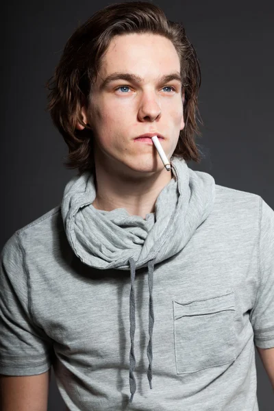 Красивый молодой человек с каштановыми длинными волосами в серой рубашке, изолированной на сером фоне. Курю сигарету. Модный студийный снимок. Экспрессивное лицо . — стоковое фото