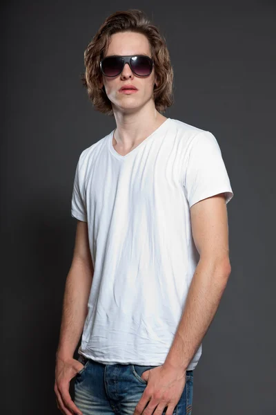 Bello giovanotto con capelli lunghi marroni e occhiali da sole isolati su sfondo grigio. Studio di moda girato . — Foto Stock