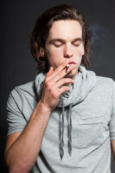 stock image Handsome young man with brown long hair wearing grey shirt isolated on grey background. Smoking cigarette. Fashion studio shot. Expressive face.