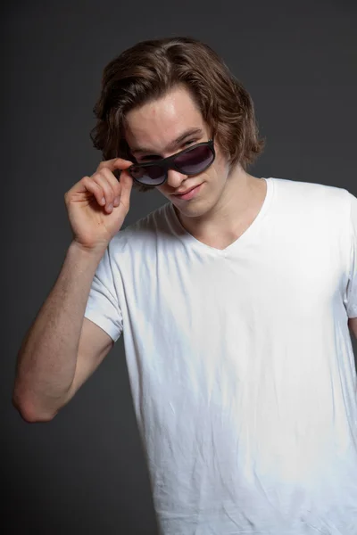 Knappe jonge man met bruine lange haren en zonnebril geïsoleerd op een grijze achtergrond. mode studio opname. — Stockfoto