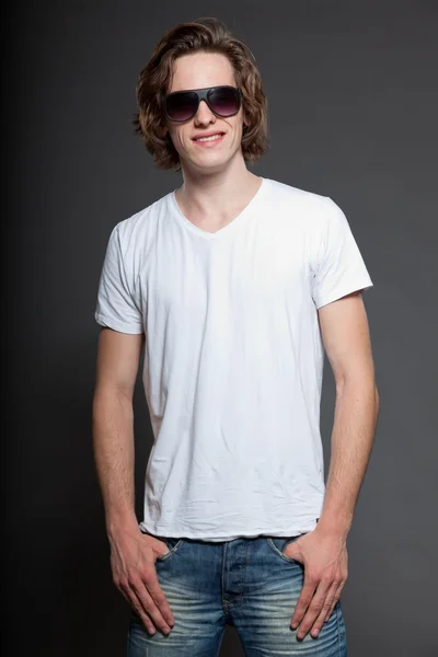 Przystojny, młody człowiek z brązowe długie włosy i okulary na białym tle na szarym tle. strzał studio mody. — Zdjęcie stockowe