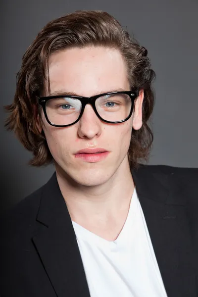 Handsome young man with brown long hair and wearing white shirt and blue jacket and retro glasses isolated on grey background. Fashion studio shot. Expressive face. — Stock Photo, Image