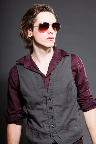 Pohledný mladý muž s hnědé dlouhé vlasy izolovaných na šedém pozadí. sluneční brýle. módní studio zastřelen. výraznou tvář. — Stock fotografie