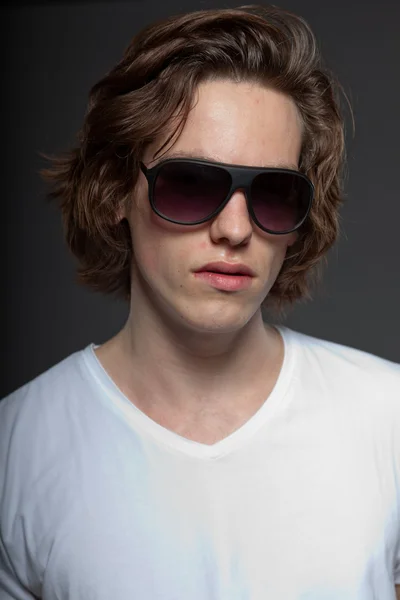Pohledný mladý muž s hnědé dlouhé vlasy a sluneční brýle, izolované na šedém pozadí. módní studio zastřelil. — Stock fotografie