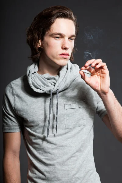 stock image Handsome young man with brown long hair wearing grey shirt isolated on grey background. Smoking cigarette. Fashion studio shot. Expressive face.