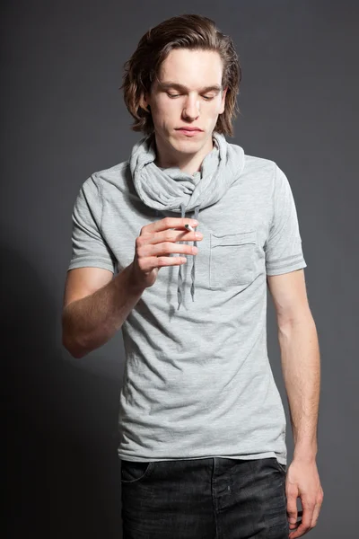 Pohledný mladý muž s hnědé dlouhé vlasy nosí šedé tričko izolovaných na šedém pozadí. kouření cigaret. módní studio zastřelen. výraznou tvář. — Stock fotografie
