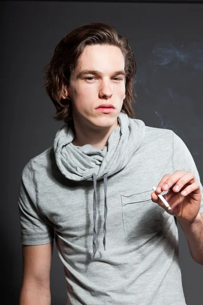 Beau jeune homme aux cheveux longs bruns portant une chemise grise isolée sur fond gris. Fumer une cigarette. Studio de mode tourné. Visage expressif . — Photo