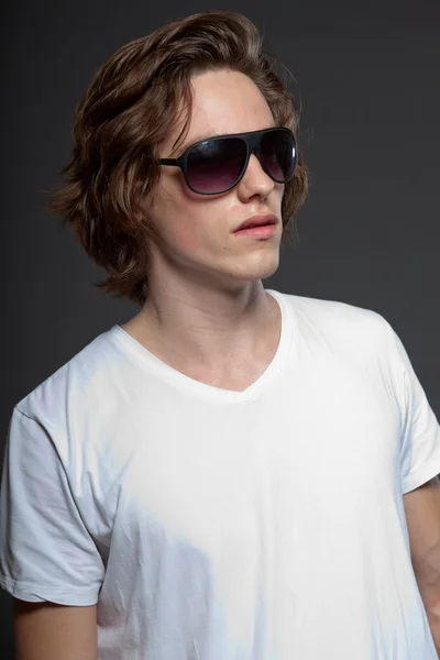 Schöner junger Mann mit braunen langen Haaren und Sonnenbrille auf grauem Hintergrund. Modestudio erschossen. — Stockfoto