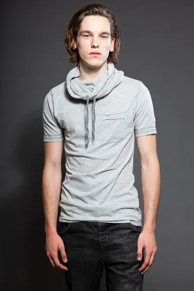 Pohledný mladý muž s hnědé dlouhé vlasy nosí šedé tričko izolovaných na šedém pozadí. módní studio zastřelen. výraznou tvář. — Stock fotografie