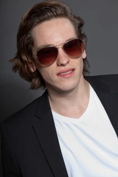 Красивый молодой человек с каштановыми длинными волосами и в белой рубашке и голубой куртке и солнечных очках, изолированных на сером фоне. Модный студийный снимок. Экспрессивное лицо . — стоковое фото