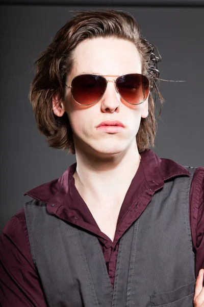 Bonito jovem com cabelo longo marrom isolado em fundo cinza. A usar óculos de sol. Estúdio de moda. Cara expressiva . — Fotografia de Stock