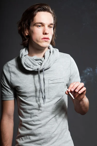 Pohledný mladý muž s hnědé dlouhé vlasy nosí šedé tričko izolovaných na šedém pozadí. kouření cigaret. módní studio zastřelen. výraznou tvář. — Stock fotografie