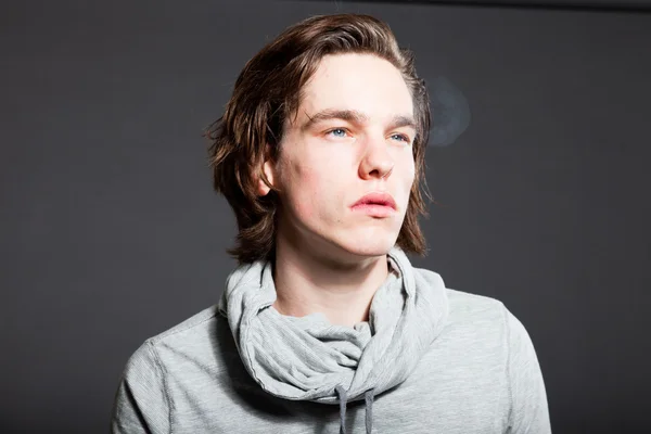 Handsome young man with brown long hair wearing grey shirt isolated on grey background. Smoking cigarette. Fashion studio shot. Expressive face. — Stock Photo, Image