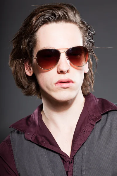 Όμορφος νεαρός άνδρας με καστανά μακριά μαλλιά που απομονώνονται σε γκρι φόντο. φορώντας γυαλιά ηλίου. στούντιο μόδας πυροβόλησε. εκφραστικό πρόσωπο. — Φωτογραφία Αρχείου