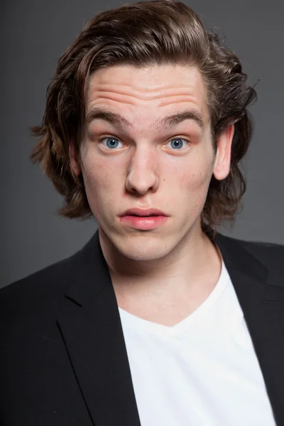 Schöner junger Mann mit braunen langen Haaren und weißem Hemd und blauer Jacke auf grauem Hintergrund. Modestudio gedreht. Gesichtsausdruck. — Stockfoto