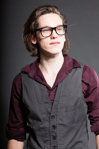 Schöner junger Mann mit braunen langen Haaren auf grauem Hintergrund. trägt eine schwarze Retro-Brille. Modestudio gedreht. Gesichtsausdruck. — Stockfoto