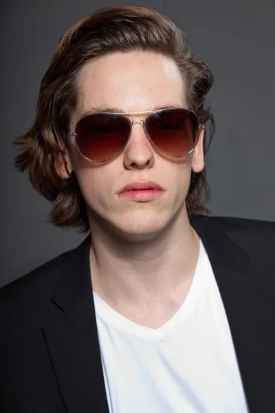 Handsome young man with brown long hair and wearing white shirt and blue jacket and sunglasses isolated on grey background. Fashion studio shot. Expressive face. — Stock Photo, Image