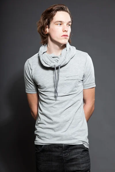 Pohledný mladý muž s hnědé dlouhé vlasy nosí šedé tričko izolovaných na šedém pozadí. módní studio zastřelen. výraznou tvář. — Stock fotografie