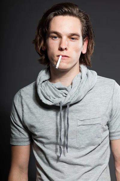 Όμορφος νεαρός άνδρας με καστανά μακριά μαλλιά φορώντας γκρι πουκάμισο που απομονώνονται σε γκρι φόντο. το κάπνισμα τσιγάρων. στούντιο μόδας πυροβόλησε. εκφραστικό πρόσωπο. — Φωτογραφία Αρχείου