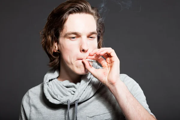 Schöner junger Mann mit braunen langen Haaren, graues Hemd auf grauem Hintergrund. Zigarette rauchend. Modestudio gedreht. Gesichtsausdruck. — Stockfoto