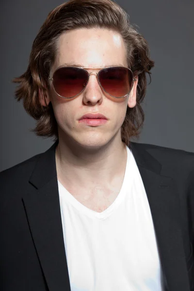Csinos, fiatal férfi barna hosszú haj és a fehér inget és kék kabát, és elszigetelt szürke háttér napszemüveg viselése. divat stúdió felvétel. kifejező arc. — Stock Fotó