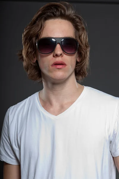 Pohledný mladý muž s hnědé dlouhé vlasy a sluneční brýle, izolované na šedém pozadí. módní studio zastřelil. — Stock fotografie