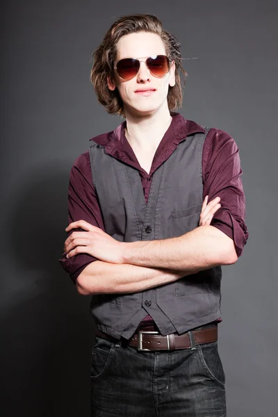 Schöner junger Mann mit braunen langen Haaren auf grauem Hintergrund. mit Sonnenbrille. Modestudio gedreht. Gesichtsausdruck. — Stockfoto
