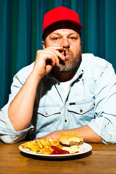Άνθρωπος με γένια τρώνε γεύμα fast food. απολαμβάνοντας τις πατάτες τηγανιτές και ένα χάμπουργκερ. καπνίζοντας ένα πούρο. φορτηγατζή με το κόκκινο σκουφί. — Φωτογραφία Αρχείου