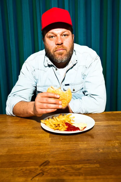 Człowiek z brodą, jedzenie fast food posiłek. korzystających z hamburger i frytki. Czapki (tirówki) z Czerwonym Kapturku. — Zdjęcie stockowe