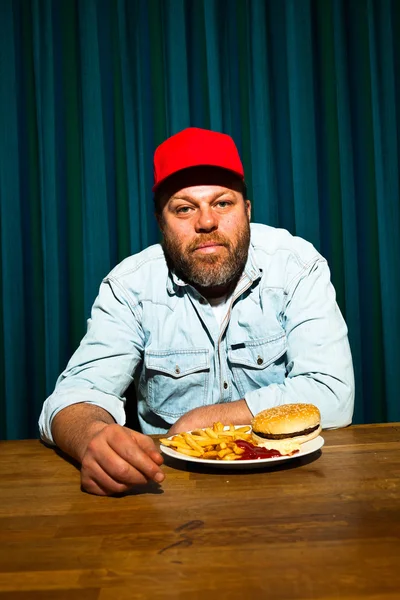 Muž s plnovousem, fast food jídlo. těší, hranolky a hamburger. Trucker s červeným víčkem. — Stock fotografie