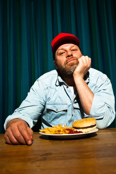 Mann mit Bart isst Fast-Food-Essen. Pommes und einen Hamburger genießen. Trucker mit roter Mütze. — Stockfoto
