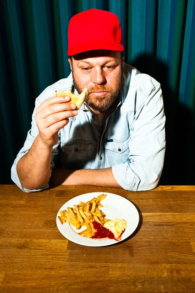 Человек с бородой ест фаст-фуд. Наслаждаюсь картошкой фри и гамбургером. дальнобойщик в красной кепке . — стоковое фото