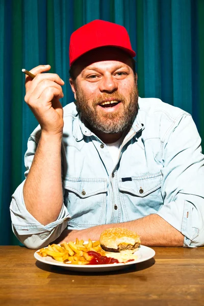 Un uomo con la barba che mangia fast food. Mi godo patatine fritte e un hamburger. Fumo un sigaro. Camionista con cappuccio rosso . — Foto Stock