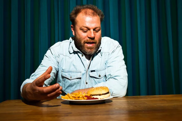 Homem com barba a comer comida rápida. Gozando de batatas fritas e um hambúrguer . — Fotografia de Stock