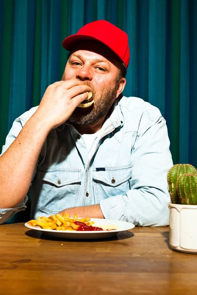 Muž s plnovousem, fast food jídlo. těší, hranolky a hamburger. Trucker s červeným víčkem. — Stock fotografie