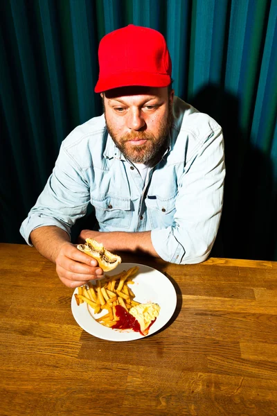 Hombre con barba comiendo comida rápida. Disfrutando de papas fritas y una hamburguesa. Camionero con gorra roja . — Foto de Stock