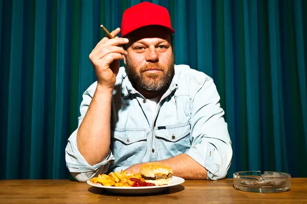 Man met baard eten van fast-food maaltijd. genieten van Franse frietjes en een hamburger. roken een sigaar. Trucker met rode dop. — Stockfoto