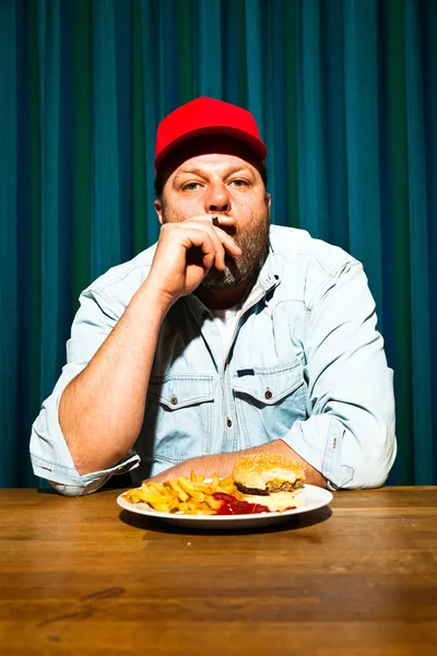 Homme à la barbe mangeant un repas de restauration rapide. Savourer des frites et un hamburger. Fumer un cigare. Camionnette avec capuchon rouge . — Photo