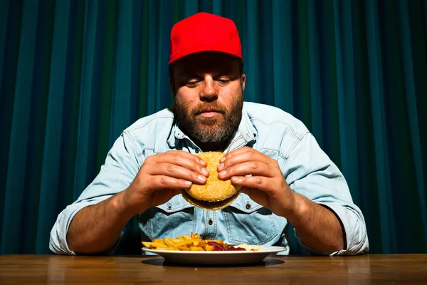 Człowiek z brodą, jedzenie fast food posiłek. korzystających z hamburger i frytki. Czapki (tirówki) z Czerwonym Kapturku. — Zdjęcie stockowe