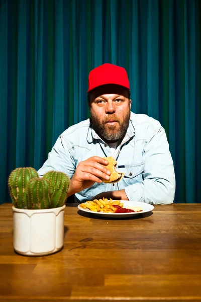 Homem com barba a comer comida rápida. A desfrutar de batatas fritas e de um hambúrguer. Camionista com tampa vermelha . — Fotografia de Stock
