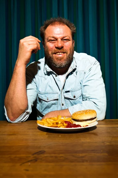 Homem com barba a comer comida rápida. Gozando de batatas fritas e um hambúrguer . — Fotografia de Stock