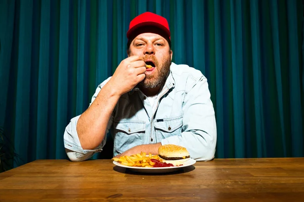Hombre con barba comiendo comida rápida. Disfrutando de papas fritas y una hamburguesa. Camionero con gorra roja . — Foto de Stock