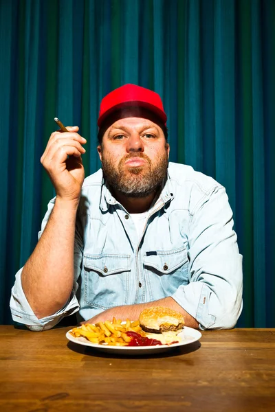 Hombre con barba comiendo comida rápida. Disfrutando de papas fritas y una hamburguesa. Fumar un cigarro. Camionero con gorra roja . —  Fotos de Stock